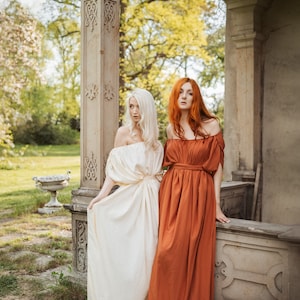 Puede incluir: Dos mujeres con vestidos largos y fluidos se encuentran en un jard&iacute;n. Una mujer tiene el pelo largo y rubio y lleva un vestido blanco. La otra mujer tiene el pelo largo y rojo y lleva un vestido color &oacute;xido. Est&aacute;n de pie frente a un pilar de piedra.