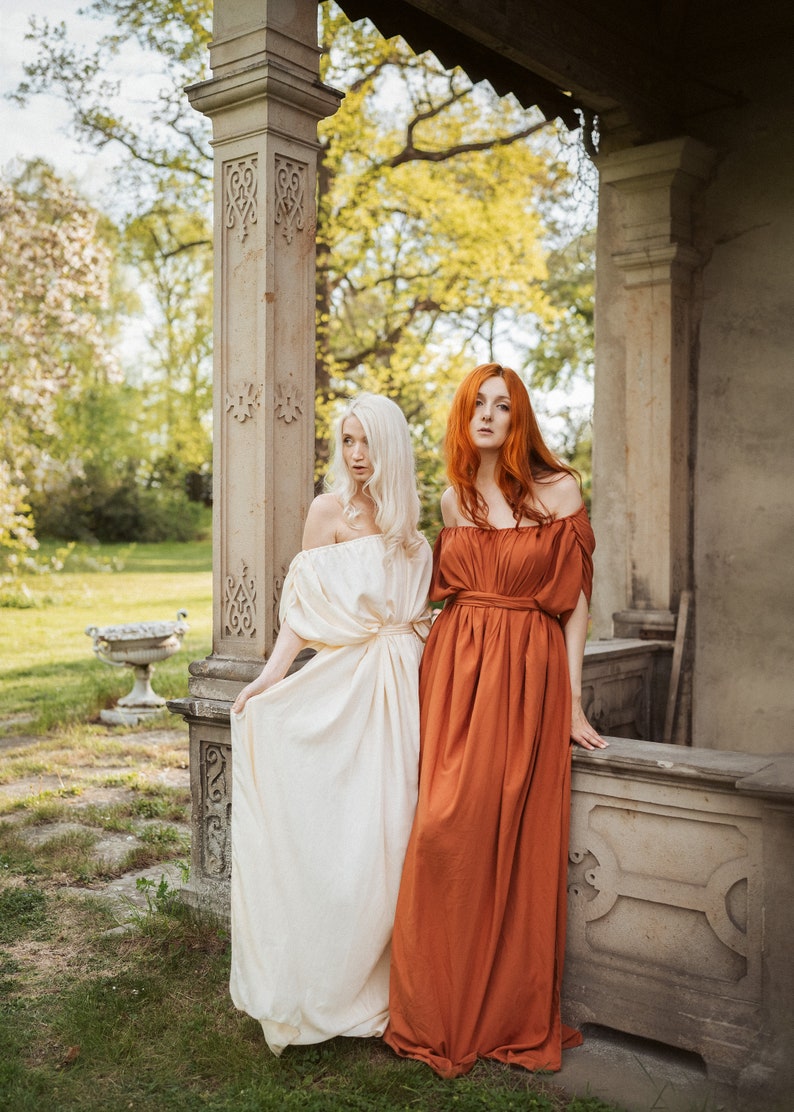 Puede incluir: Dos mujeres con vestidos largos y fluidos se encuentran en un jard&iacute;n. Una mujer tiene el pelo largo y rubio y lleva un vestido blanco. La otra mujer tiene el pelo largo y rojo y lleva un vestido color &oacute;xido. Est&aacute;n de pie frente a un pilar de piedra.