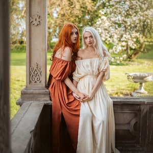 Puede incluir: Dos mujeres con vestidos largos, una con un vestido color &oacute;xido y la otra con un vestido blanco, est&aacute;n sentadas en un borde de piedra. Est&aacute;n mirando a la c&aacute;mara.