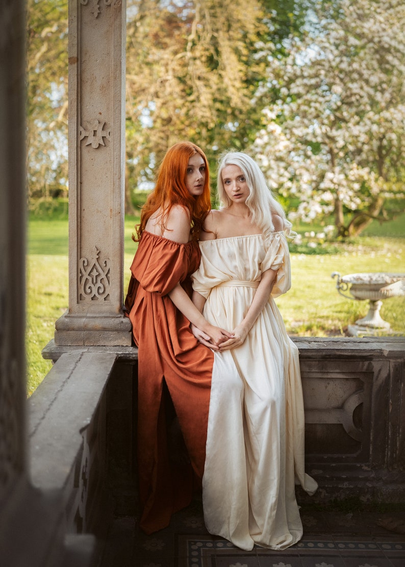 Puede incluir: Dos mujeres con vestidos largos, una con un vestido color &oacute;xido y la otra con un vestido blanco, est&aacute;n sentadas en un borde de piedra. Est&aacute;n mirando a la c&aacute;mara.