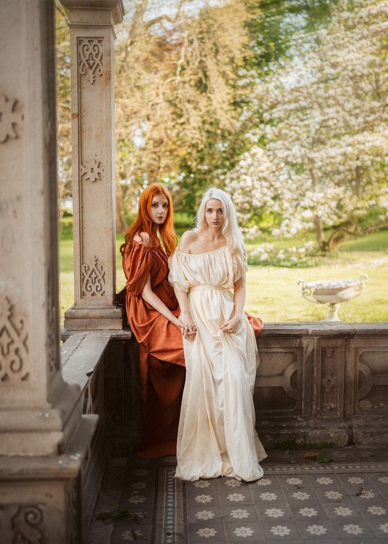 Puede incluir: Dos mujeres con vestidos largos se paran en un borde de piedra. La mujer de la izquierda lleva un vestido color &oacute;xido y la mujer de la derecha lleva un vestido blanco. Ambas miran a la c&aacute;mara.