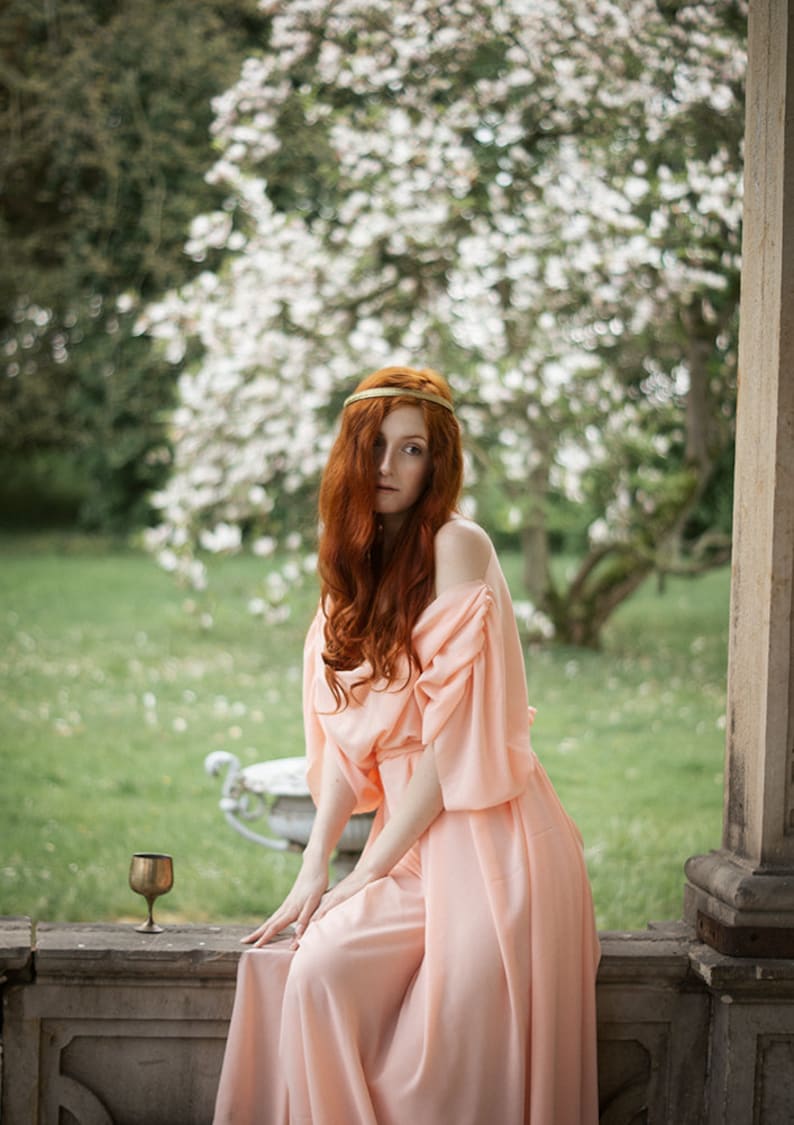 May include: A woman with long, red hair wearing a peach-colored dress and a gold headband sits on a stone ledge. She is looking to the side and there is a small gold goblet on the ledge in front of her.