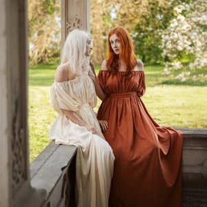 Puede incluir: Dos mujeres con vestidos largos se encuentran en un jard&iacute;n. Una mujer lleva un vestido blanco y la otra lleva un vestido color &oacute;xido. Est&aacute;n de pie cerca de un pilar de piedra.