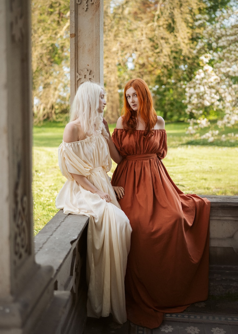 Puede incluir: Dos mujeres con vestidos largos se encuentran en un jard&iacute;n. Una mujer lleva un vestido blanco y la otra lleva un vestido color &oacute;xido. Est&aacute;n de pie cerca de un pilar de piedra.