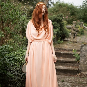 May include: A woman with long, red hair wearing a peach-colored, flowing dress stands on a stone path in a garden setting.
