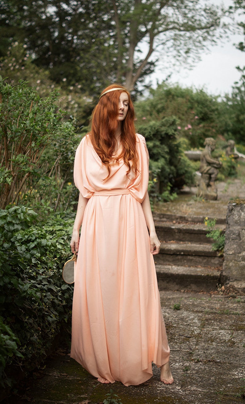May include: A woman with long, red hair wearing a peach-colored, flowing dress stands on a stone path in a garden setting.