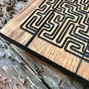 May include: A wooden board with a black geometric pattern carved into the surface. The board is made of light brown wood and has a dark brown stain.