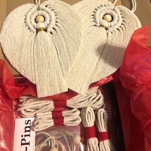 May include: Two white heart-shaped macrame wall hangings with wooden beads and tassels. The hangings are displayed on a red tissue paper background with a comb and white string.