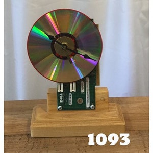 May include: A clock made from a computer circuit board and a CD. The clock is mounted on a wooden base. The clock face is a CD with a rainbow iridescent finish. The clock hands are black.