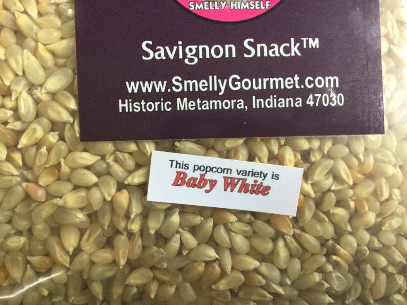 May include: A close-up of a bag of white popcorn kernels. The label reads "Savignon Snack&trade;" and "www.SmellyGourmet.com". A smaller label reads "This popcorn variety is Baby White".