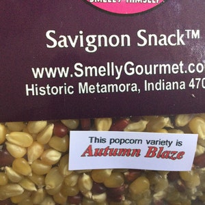 May include: A close-up of a bag of popcorn kernels. The kernels are a mix of yellow, red, and white. The bag has a label that says "Savignon Snack&trade;" and "www.SmellyGourmet.com". The label also says "Historic Metamora, Indiana 47030". A smaller label on the bag says "This popcorn variety is Autumn Blaze".