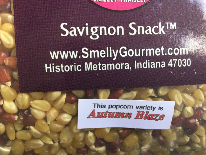 May include: A close-up of a bag of popcorn kernels. The kernels are a mix of yellow, red, and white. The bag has a label that says "Savignon Snack&trade;" and "www.SmellyGourmet.com". The label also says "Historic Metamora, Indiana 47030". A smaller label on the bag says "This popcorn variety is Autumn Blaze".