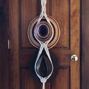 May include: A white macrame hat rack hanging on a wooden door with two hats hanging from it. The top hat is a multi-colored straw hat and the bottom hat is a brown felt hat.