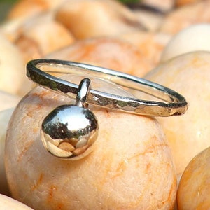 Puede incluir: Un anillo de plata con un pequeño charm redondo colgando. El anillo está sobre un lecho de piedras lisas de color marrón claro.