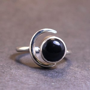 Pode incluir: Anel de prata com uma pedra preciosa preta grande e redonda, definida em um design em forma de crescente. O anel tem um acabamento polido e um estilo simples e elegante, adequado para uso diário ou ocasiões especiais.