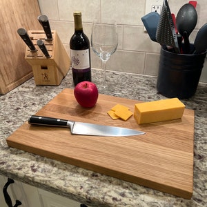 May include: A wooden cutting board with a stainless steel knife, red apple, and cheese. A bottle of red wine and a wine glass are also present. A wooden knife block with knives is in the background.