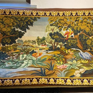 Puede incluir: Un tapiz detallado que representa un paisaje sereno con un río, árboles y aves. La obra de arte presenta un borde marrón y dorado. El tapiz se exhibe sobre un sofá de cuero marrón.