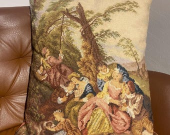 May include: Square tapestry pillow featuring a pastoral scene with figures in period clothing. The pillow has a beige background with a detailed woven design in shades of brown, green, pink, and blue. The pillow is placed on a brown leather couch.