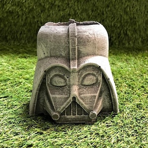 May include: A grey concrete planter shaped like Darth Vader's head. The planter has a textured surface and a detailed design of Darth Vader's face.