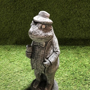 May include: A grey concrete garden statue of a frog wearing a hat and a suit holding a glass. The frog is standing on a base.