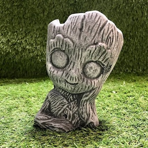May include: A grey concrete planter shaped like the character Groot from the Guardians of the Galaxy movie franchise. The planter has a textured surface and a small opening at the top for planting.