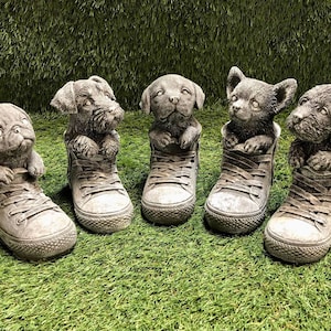May include: Five grey concrete dog figurines, each wearing a grey and white trainer. The dogs are all different breeds and are looking up at the camera.