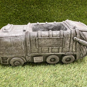 May include: A grey concrete planter shaped like a garbage truck. The planter has a rectangular opening on top for planting.