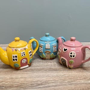 May include: Three ceramic teapots shaped like houses. The teapots are yellow, blue and pink. Each teapot has windows and a door.