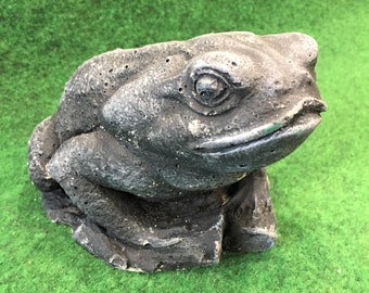Puede incluir: Una figurita de rana de piedra gris sentada en una roca. La rana tiene una superficie texturizada y una cara detallada.