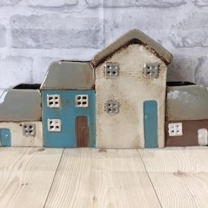 May include: A ceramic planter shaped like a row of houses with grey roofs, white walls, and blue and brown accents. The planter has three separate compartments for plants.