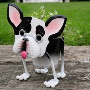 May include: A white and black metal planter shaped like a French bulldog with a pink tongue sticking out. The planter has large, round eyes and is standing on four legs. The planter is filled with green plants and white flowers.