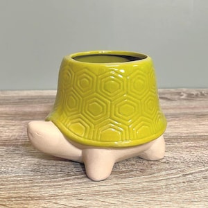 May include: A ceramic turtle-shaped planter in a vibrant yellow-green colour. The turtle's shell features a textured hexagonal pattern, while the base and feet are a neutral beige. The planter is designed to hold small plants or succulents.