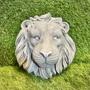 May include: A grey stone lion head sculpture with a detailed mane and facial features. The sculpture is set against a backdrop of green artificial grass. This decorative piece is suitable for garden or home decor. The sculpture is made of stone.