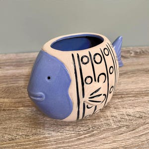 May include: A ceramic fish-shaped planter. The head and tail are blue, and the body is cream-coloured with black lines and circles. The interior is blue. The planter is on a wooden surface.