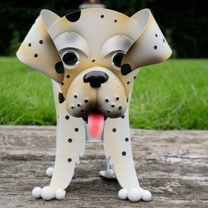 May include: A metal dog planter, cream-coloured with black spots, resembling a Dalmatian. It features large eyes, a black nose, and a pink tongue. The planter is designed to hold plants and is placed on a wooden surface.