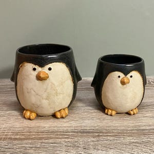 May include: Two ceramic penguin planters, one larger than the other, with black and white painted details. The smaller planter is in front of the larger planter.