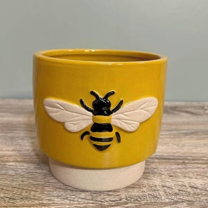 May include: A yellow ceramic plant pot featuring a raised bee design. The bee has black and yellow stripes with white wings. The pot sits on a white base, suitable for small plants or succulents.