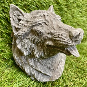 May include: A grey, textured wolf head sculpture with an open mouth and visible tongue. The sculpture has detailed fur and is set against a backdrop of green grass. This decorative piece is likely for garden or home decor.