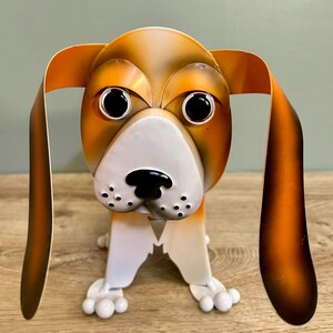 May include: A metal dog sculpture in brown and white. The dog has large black eyes, long floppy ears, and a white snout with black spots. The sculpture stands on four white paws. A decorative item.
