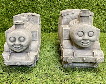 May include: Two gray concrete train sculptures with smiling faces, set against a backdrop of green grass. The trains have circular eyes, a round nose, and a chimney. The sculptures are likely garden decor.