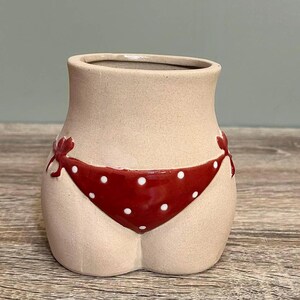May include: A beige ceramic vase shaped like a torso, featuring a red polka-dot bikini design. The bikini has white polka dots and red bows. The vase has a smooth, matte finish, and is designed to hold flowers.