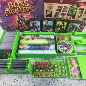Pode incluir: Um jogo de tabuleiro Boss Fighters com uma caixa grande, quatro baralhos de personagens e vários componentes organizados em tabuleiros de plástico verde brilhante. O conjunto inclui tapetes de jogo enrolados e fichas circulares.