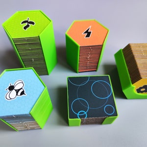 K&ouml;nnte beinhalten: F&uuml;nf Stapel sechseckiger Spielkarten mit gr&uuml;nen, orangefarbenen, blauen und schwarzen Kartenfl&auml;chen. Die Karten sind in einem gr&uuml;nen Rahmen gestapelt. Die Kartenfl&auml;chen zeigen ein Blatt, einen Blitz, eine Biene und ein Muster aus Kreisen.