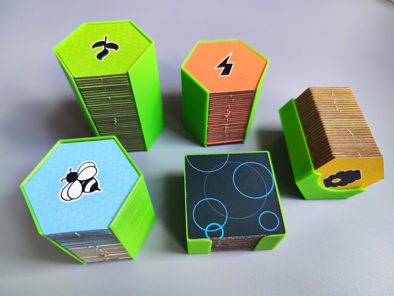 K&ouml;nnte beinhalten: F&uuml;nf Stapel sechseckiger Spielkarten mit gr&uuml;nen, orangefarbenen, blauen und schwarzen Kartenfl&auml;chen. Die Karten sind in einem gr&uuml;nen Rahmen gestapelt. Die Kartenfl&auml;chen zeigen ein Blatt, einen Blitz, eine Biene und ein Muster aus Kreisen.