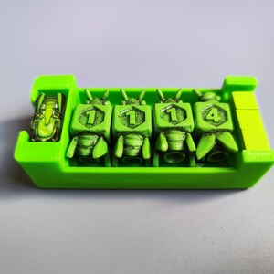K&ouml;nnte beinhalten: Ein gr&uuml;nes Plastiktablett mit vier gr&uuml;nen W&uuml;rfeln. Jeder W&uuml;rfel hat eine Zahl darauf. Die Zahlen sind 1, 1, 1 und 4. Es befindet sich ein kleines gr&uuml;nes und gelbes Objekt im Tablett.