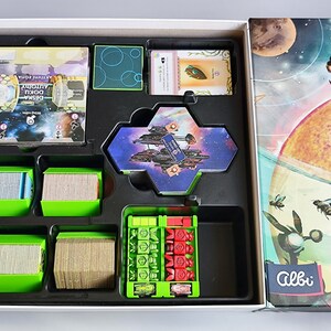 K&ouml;nnte beinhalten: Das Innere einer Brettspielbox mit gr&uuml;nen und schwarzen F&auml;chern. Die F&auml;cher enthalten verschiedene Spielteile, darunter Karten, Spielsteine und eine sechseckige Kachel mit einem Raumschiff darauf. Der Deckel der Box zeigt eine farbenfrohe Illustration eines Raumschiffs und Planeten.