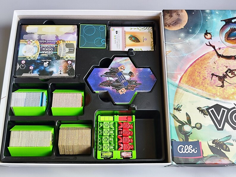 K&ouml;nnte beinhalten: Das Innere einer Brettspielbox mit gr&uuml;nen und schwarzen F&auml;chern. Die F&auml;cher enthalten verschiedene Spielteile, darunter Karten, Spielsteine und eine sechseckige Kachel mit einem Raumschiff darauf. Der Deckel der Box zeigt eine farbenfrohe Illustration eines Raumschiffs und Planeten.