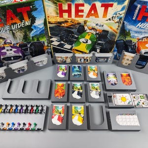 Puede incluir: Componentes de juego de mesa con coches de carreras y cartas coloridas. Las cajas del juego, con la palabra "HEAT", son visibles en el fondo. Varias piezas y soportes de cartas están dispuestos sobre una superficie gris.