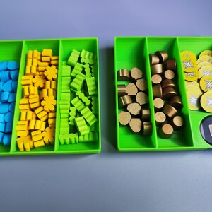 K&ouml;nnte beinhalten: Zwei gr&uuml;ne Kunststoffschalen mit F&auml;chern, gef&uuml;llt mit bunten Spielsteinen. Eine Schale hat blaue, gelbe und gr&uuml;ne Steine. Die andere Schale hat goldene Steine und runde Spielmarken mit einem gelben und wei&szlig;en Design.
