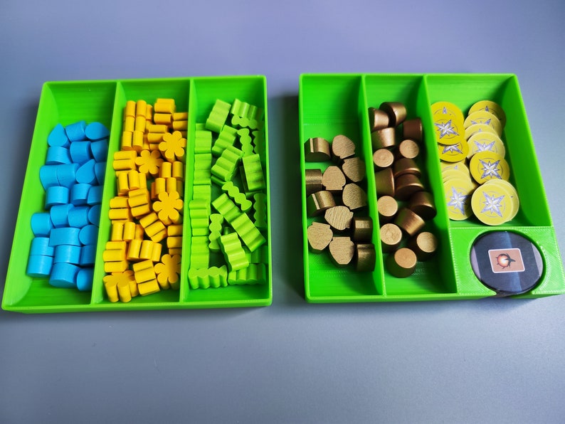 K&ouml;nnte beinhalten: Zwei gr&uuml;ne Kunststoffschalen mit F&auml;chern, gef&uuml;llt mit bunten Spielsteinen. Eine Schale hat blaue, gelbe und gr&uuml;ne Steine. Die andere Schale hat goldene Steine und runde Spielmarken mit einem gelben und wei&szlig;en Design.
